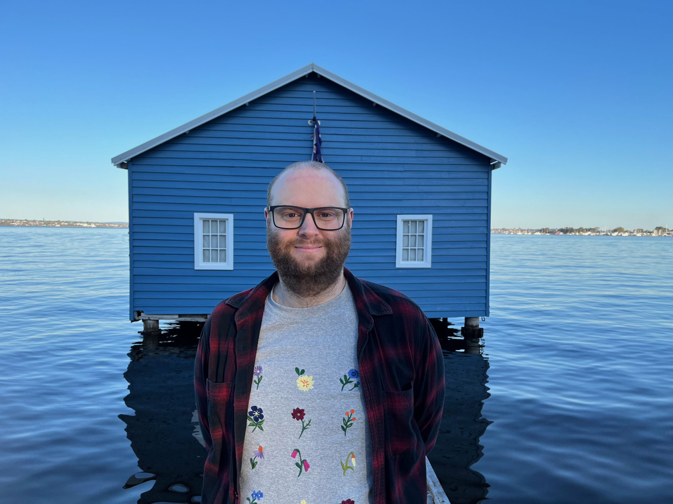 Dr James Lloyd Research Fellow at the University of Western Australia and the ARC Centre of Excellence in Plants for Space stands in front of a blue boat house in perth
